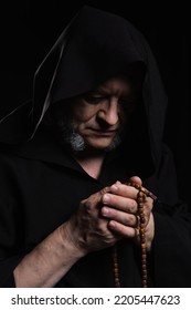 Medieval Priest In Hooded Robe Praying With Rosary Beads Isolated On Black