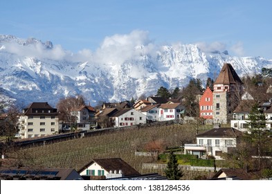 Medieval Red House And Vineyards In The Capital Of Liechtenstein In The City Of Vaduz On The Background Of Residential Buildings, Blue Sky And Snow-capped Mountains