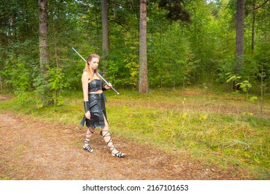 Medieval Robber Girl Wearing Leather Lorica, Bracer, Rerebrace And Skirt With Big Falchion On Her Shoulder Walks In The Summer Wood