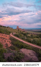 Medieval towers stand majestically on a rocky hill, overlooking a winding dirt road and vast open landscape under a pastel-colored sky