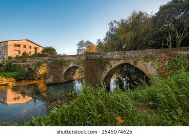 The medieval Traba Bridge spans a calm river, surrounded by lush greenery and ivy-covered stone, with an old building reflecting in the water under a serene sky. - Powered by Shutterstock