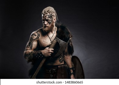 Средневековый воин-берсерк викинг с татуировкой на коже, рыжей бородой и косами в волосах с топором и щитом атакует врага. Концептуальная историческая фотография