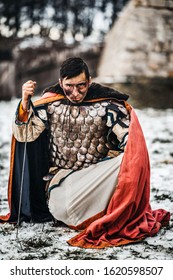 A Medieval Warrior In Chain Mail Armor And Mantle Kneeling And Holds His Hand To Sword After Battle. Background Of Winter Forest And Snow. Man Looking Into The Camera.