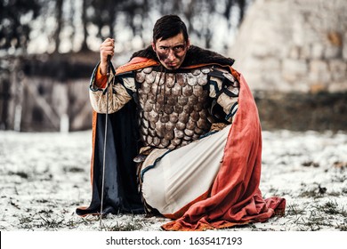 A Medieval Warrior In Chain Mail Armor And Mantle Kneeling And Holds His Hand To Sword After Battle. Background Of Winter Forest And Snow. Man Looking Into The Camera.