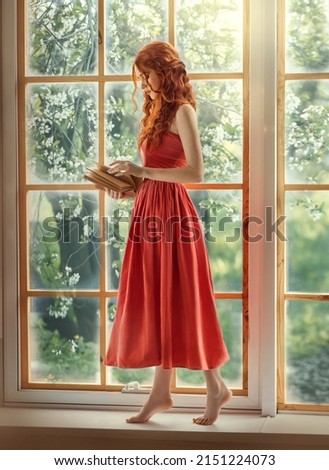 Medieval woman princess holds in hands reads book. Red-haired dreamlike girl stands on window barefoot, view summer green garden trees. Red vintage dress, ball gown. Long red curly hair, pale skin