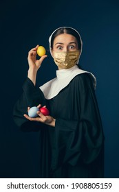 Medieval Young Woman As A Nun In Vintage Clothing And Golden Face Mask With Easter Eggs On Dark Blue Background. Concept Of Comparison Of Eras, Fashion, Ad, Healthcare. Royal Person Protected From