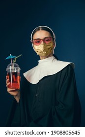 Medieval Young Woman As A Nun In Vintage Clothing And Golden Face Mask With Cocktail On Dark Blue Background. Concept Of Comparison Of Eras, Fashion, Ad, Healthcare. Royal Person Protected From Covid.