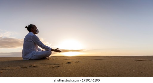 meditating man in medical mask in lotus pose on the beach, male wearing surgical face mask and white yoga clothes with man bun top knot hairstyle sitting in a meditation pose on an empty beach sunrise - Powered by Shutterstock