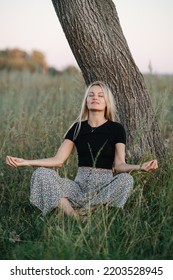 Meditating Under A Tree Blond Woman Sitting On Grass Cross-legged. She Is Wearing Skirt Pants And Black Shirt. Her Eyes Closed. Low Angle.