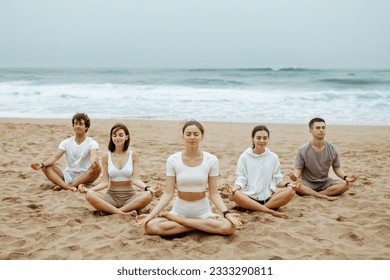 Meditation class outoors. Group of fit young people practicing yoga on the beach, meditating with closed eyes in lotus position and enjoying morning training on ocean shore - Powered by Shutterstock