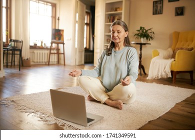 Meditation, self-awareness, healthy body and mind. Casually dressed mature woman sitting on floor in front of laptop, legs crossed, closing eyes, meditating, listening to calm music or affirmations