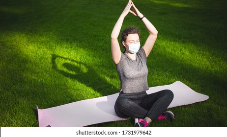 Meditation Yoga Woman In Medical Protective Mask On Green Grass Meditating On Sunset Sunrise In Park, Sitting In Lotus Position.Practicing Yoga Workout Outdoor.Active Life During Coronavirus