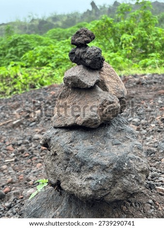 A meditative stack of volcanic rocks, known as a cairn, carefully balanced on a rugged gravel path. Set against a lush green forest backdrop under a misty sky, this image symbolizes balance, stability, and a spiritual connection with nature. Perfect for concepts of mindfulness, zen, and outdoor exploration.