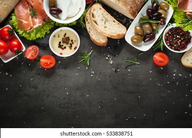 Mediterranean Food Background. Ciabatta Bread Jamon Serrano Tomatoes Herbs And Olives On Black Slate Board. Top View