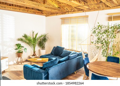 Mediterranean Interior Design Of Beautiful House, Living Room With Big Sofa And Home Green Plants. Cozy Indoor Place With Wooden Elements And Bamboo Blinds Transmitting The Rays Of The Sun.