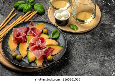 mediterranean salad with prosciutto or jamon and green basil leaves and Cantaloupe melon on dark table, traditional Spanish and Italian appetizer served with wholemeal grissini, top view.