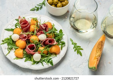 mediterranean salad with prosciutto or jamon, mozzarella and green basil leaves and Cantaloupe melon on white table, traditional Spanish and Italian appetizer served with wholemeal grissini, top view,
