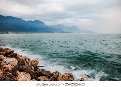 Mediterranean Sea. Waves crash against rocky shores, with a backdrop of misty mountains under an overcast sky. The expansive sea meets the horizon, creating a tranquil and scenic view. - Powered by Shutterstock