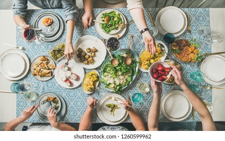 Mediterranean Style Dinner. Flat-lay Of Table With Salads, Starters, Pastries Over Blue Table Cloth With Hands Holding Drinks, Sharing Food, Top View. Holiday Gathering And Vegetarian Party Concept