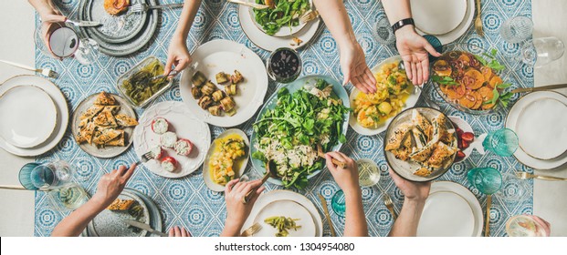 Mediterranean Style Dinner. Flat-lay Of Table With Salads, Starters, Pastries Over Blue Table Cloth With Hands Holding Drinks, Sharing Food, Top View, Wide Composition. Holiday Gathering Party Concept