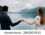 A medium back view shot capturing a young romantic couple standing together and looking at a beautiful alpine scenery, famous location in Switzerland. A man in a dark sweater and a woman in a light summer dress are holding hands or hugging while enjoying a peaceful holiday on the wooden pier platform next to the water. In the background, a historic European castle or palace stands on the edge of a small peninsula surrounded by lush green trees and pristine nature. The calm turquoise water of Lake Brienz or Lake Thun reflects the dramatic cloudy sky, creating an incredibly idyllic, misty, and moody atmosphere typical for the Bernese Oberland region near Interlaken. A large white passenger cruise ship or tourist ferry boat floats slowly on the deep mountain lake, carrying international travelers who explore the stunning Swiss landscape during their summer vacation trip. High mountain slopes covered with green forests rise into the thick low clouds, adding a cinematic and spectacular wilderness look to this professional lifestyle photography session. This beautiful composition perfectly represents high-end destination wedding travel, engagement planning, family honeymoon getaways, and outdoor tourism in western Europe. The soft natural daylight and authentic emotions of the lovers create a sophisticated mood, making it an excellent stock image for bridal magazines, travel agencies, luxury hospitality brands, or editorial content. Every element, from the clean architectural details of the distant lakeside estate to the gentle ripples on the water surface and the couple