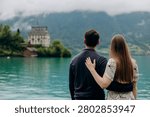 A medium back view shot capturing a young romantic couple standing together and looking at a beautiful alpine scenery, famous location in Switzerland. A man in a dark sweater and a woman in a light summer dress are holding hands or hugging while enjoying a peaceful holiday on the wooden pier platform next to the water. In the background, a historic European castle or palace stands on the edge of a small peninsula surrounded by lush green trees and pristine nature. The calm turquoise water of Lake Brienz or Lake Thun reflects the dramatic cloudy sky, creating an incredibly idyllic, misty, and moody atmosphere typical for the Bernese Oberland region near Interlaken. A large white passenger cruise ship or tourist ferry boat floats slowly on the deep mountain lake, carrying international travelers who explore the stunning Swiss landscape during their summer vacation trip. High mountain slopes covered with green forests rise into the thick low clouds, adding a cinematic and spectacular wilderness look to this professional lifestyle photography session. This beautiful composition perfectly represents high-end destination wedding travel, engagement planning, family honeymoon getaways, and outdoor tourism in western Europe. The soft natural daylight and authentic emotions of the lovers create a sophisticated mood, making it an excellent stock image for bridal magazines, travel agencies, luxury hospitality brands, or editorial content. Every element, from the clean architectural details of the distant lakeside estate to the gentle ripples on the water surface and the couple