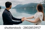 A medium back view shot capturing a young romantic couple standing together and looking at a beautiful alpine scenery, famous location in Switzerland. A man in a dark sweater and a woman in a light summer dress are holding hands or hugging while enjoying a peaceful holiday on the wooden pier platform next to the water. In the background, a historic European castle or palace stands on the edge of a small peninsula surrounded by lush green trees and pristine nature. The calm turquoise water of Lake Brienz or Lake Thun reflects the dramatic cloudy sky, creating an incredibly idyllic, misty, and moody atmosphere typical for the Bernese Oberland region near Interlaken. A large white passenger cruise ship or tourist ferry boat floats slowly on the deep mountain lake, carrying international travelers who explore the stunning Swiss landscape during their summer vacation trip. High mountain slopes covered with green forests rise into the thick low clouds, adding a cinematic and spectacular wilderness look to this professional lifestyle photography session. This beautiful composition perfectly represents high-end destination wedding travel, engagement planning, family honeymoon getaways, and outdoor tourism in western Europe. The soft natural daylight and authentic emotions of the lovers create a sophisticated mood, making it an excellent stock image for bridal magazines, travel agencies, luxury hospitality brands, or editorial content. Every element, from the clean architectural details of the distant lakeside estate to the gentle ripples on the water surface and the couple