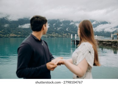 A medium back view shot capturing a young romantic couple standing together and looking at a beautiful alpine scenery, famous location in Switzerland. A man in a dark sweater and a woman in a light summer dress are holding hands or hugging while enjoying a peaceful holiday on the wooden pier platform next to the water. In the background, a historic European castle or palace stands on the edge of a small peninsula surrounded by lush green trees and pristine nature. The calm turquoise water of Lake Brienz or Lake Thun reflects the dramatic cloudy sky, creating an incredibly idyllic, misty, and moody atmosphere typical for the Bernese Oberland region near Interlaken. A large white passenger cruise ship or tourist ferry boat floats slowly on the deep mountain lake, carrying international travelers who explore the stunning Swiss landscape during their summer vacation trip. High mountain slopes covered with green forests rise into the thick low clouds, adding a cinematic and spectacular wilderness look to this professional lifestyle photography session. This beautiful composition perfectly represents high-end destination wedding travel, engagement planning, family honeymoon getaways, and outdoor tourism in western Europe. The soft natural daylight and authentic emotions of the lovers create a sophisticated mood, making it an excellent stock image for bridal magazines, travel agencies, luxury hospitality brands, or editorial content. Every element, from the clean architectural details of the distant lakeside estate to the gentle ripples on the water surface and the couple's intimate interaction, is captured with exceptional sharpness and artistic visual balance. An ideal digital asset for advertising romantic weekend tours, alpine cruise excursions, eco-travel concepts, or traditional relationship values in the heart of the Swiss Alps, showcasing the breathtaking natural heritage and premium holiday experience of a famous location in Switzerland during the overcast summer season.