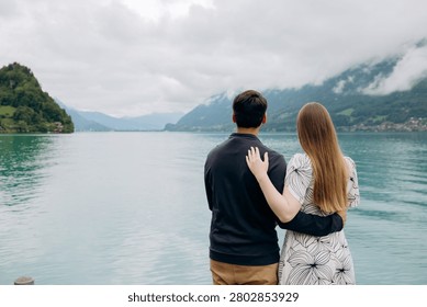 A medium back view shot capturing a young romantic couple standing together and looking at a beautiful alpine scenery, famous location in Switzerland. A man in a dark sweater and a woman in a light summer dress are holding hands or hugging while enjoying a peaceful holiday on the wooden pier platform next to the water. In the background, a historic European castle or palace stands on the edge of a small peninsula surrounded by lush green trees and pristine nature. The calm turquoise water of Lake Brienz or Lake Thun reflects the dramatic cloudy sky, creating an incredibly idyllic, misty, and moody atmosphere typical for the Bernese Oberland region near Interlaken. A large white passenger cruise ship or tourist ferry boat floats slowly on the deep mountain lake, carrying international travelers who explore the stunning Swiss landscape during their summer vacation trip. High mountain slopes covered with green forests rise into the thick low clouds, adding a cinematic and spectacular wilderness look to this professional lifestyle photography session. This beautiful composition perfectly represents high-end destination wedding travel, engagement planning, family honeymoon getaways, and outdoor tourism in western Europe. The soft natural daylight and authentic emotions of the lovers create a sophisticated mood, making it an excellent stock image for bridal magazines, travel agencies, luxury hospitality brands, or editorial content. Every element, from the clean architectural details of the distant lakeside estate to the gentle ripples on the water surface and the couple's intimate interaction, is captured with exceptional sharpness and artistic visual balance. An ideal digital asset for advertising romantic weekend tours, alpine cruise excursions, eco-travel concepts, or traditional relationship values in the heart of the Swiss Alps, showcasing the breathtaking natural heritage and premium holiday experience of a famous location in Switzerland during the overcast summer season.