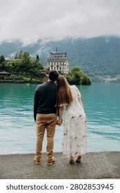 A medium back view shot capturing a young romantic couple standing together and looking at a beautiful alpine scenery, famous location in Switzerland. A man in a dark sweater and a woman in a light summer dress are holding hands or hugging while enjoying a peaceful holiday on the wooden pier platform next to the water. In the background, a historic European castle or palace stands on the edge of a small peninsula surrounded by lush green trees and pristine nature. The calm turquoise water of Lake Brienz or Lake Thun reflects the dramatic cloudy sky, creating an incredibly idyllic, misty, and moody atmosphere typical for the Bernese Oberland region near Interlaken. A large white passenger cruise ship or tourist ferry boat floats slowly on the deep mountain lake, carrying international travelers who explore the stunning Swiss landscape during their summer vacation trip. High mountain slopes covered with green forests rise into the thick low clouds, adding a cinematic and spectacular wilderness look to this professional lifestyle photography session. This beautiful composition perfectly represents high-end destination wedding travel, engagement planning, family honeymoon getaways, and outdoor tourism in western Europe. The soft natural daylight and authentic emotions of the lovers create a sophisticated mood, making it an excellent stock image for bridal magazines, travel agencies, luxury hospitality brands, or editorial content. Every element, from the clean architectural details of the distant lakeside estate to the gentle ripples on the water surface and the couple's intimate interaction, is captured with exceptional sharpness and artistic visual balance. An ideal digital asset for advertising romantic weekend tours, alpine cruise excursions, eco-travel concepts, or traditional relationship values in the heart of the Swiss Alps, showcasing the breathtaking natural heritage and premium holiday experience of a famous location in Switzerland during the overcast summer season.
