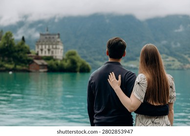A medium back view shot capturing a young romantic couple standing together and looking at a beautiful alpine scenery, famous location in Switzerland. A man in a dark sweater and a woman in a light summer dress are holding hands or hugging while enjoying a peaceful holiday on the wooden pier platform next to the water. In the background, a historic European castle or palace stands on the edge of a small peninsula surrounded by lush green trees and pristine nature. The calm turquoise water of Lake Brienz or Lake Thun reflects the dramatic cloudy sky, creating an incredibly idyllic, misty, and moody atmosphere typical for the Bernese Oberland region near Interlaken. A large white passenger cruise ship or tourist ferry boat floats slowly on the deep mountain lake, carrying international travelers who explore the stunning Swiss landscape during their summer vacation trip. High mountain slopes covered with green forests rise into the thick low clouds, adding a cinematic and spectacular wilderness look to this professional lifestyle photography session. This beautiful composition perfectly represents high-end destination wedding travel, engagement planning, family honeymoon getaways, and outdoor tourism in western Europe. The soft natural daylight and authentic emotions of the lovers create a sophisticated mood, making it an excellent stock image for bridal magazines, travel agencies, luxury hospitality brands, or editorial content. Every element, from the clean architectural details of the distant lakeside estate to the gentle ripples on the water surface and the couple's intimate interaction, is captured with exceptional sharpness and artistic visual balance. An ideal digital asset for advertising romantic weekend tours, alpine cruise excursions, eco-travel concepts, or traditional relationship values in the heart of the Swiss Alps, showcasing the breathtaking natural heritage and premium holiday experience of a famous location in Switzerland during the overcast summer season.
