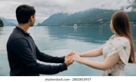 A medium back view shot capturing a young romantic couple standing together and looking at a beautiful alpine scenery, famous location in Switzerland. A man in a dark sweater and a woman in a light summer dress are holding hands or hugging while enjoying a peaceful holiday on the wooden pier platform next to the water. In the background, a historic European castle or palace stands on the edge of a small peninsula surrounded by lush green trees and pristine nature. The calm turquoise water of Lake Brienz or Lake Thun reflects the dramatic cloudy sky, creating an incredibly idyllic, misty, and moody atmosphere typical for the Bernese Oberland region near Interlaken. A large white passenger cruise ship or tourist ferry boat floats slowly on the deep mountain lake, carrying international travelers who explore the stunning Swiss landscape during their summer vacation trip. High mountain slopes covered with green forests rise into the thick low clouds, adding a cinematic and spectacular wilderness look to this professional lifestyle photography session. This beautiful composition perfectly represents high-end destination wedding travel, engagement planning, family honeymoon getaways, and outdoor tourism in western Europe. The soft natural daylight and authentic emotions of the lovers create a sophisticated mood, making it an excellent stock image for bridal magazines, travel agencies, luxury hospitality brands, or editorial content. Every element, from the clean architectural details of the distant lakeside estate to the gentle ripples on the water surface and the couple's intimate interaction, is captured with exceptional sharpness and artistic visual balance. An ideal digital asset for advertising romantic weekend tours, alpine cruise excursions, eco-travel concepts, or traditional relationship values in the heart of the Swiss Alps, showcasing the breathtaking natural heritage and premium holiday experience of a famous location in Switzerland during the overcast summer season.