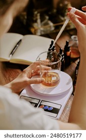 Medium close up of crystal flask with aromatic caramel liquid on kitchen scale and female hand holding plastic pipette full of essential oil