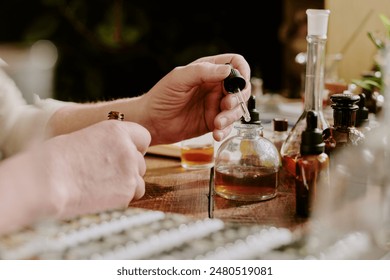Medium close up of perfumers hand holding glass pipette full of essential oil and adding it into main flask with caramel shade liquid