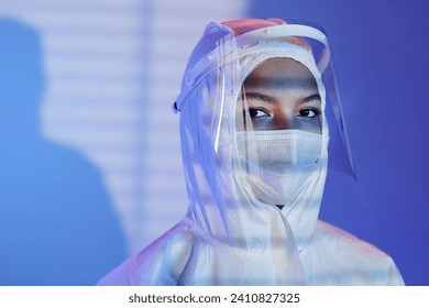 Medium close up shot female laboratory technician dressed in hazmat suit, face shield and mask looking at camera while standing at blue background with blinds shadow - Powered by Shutterstock