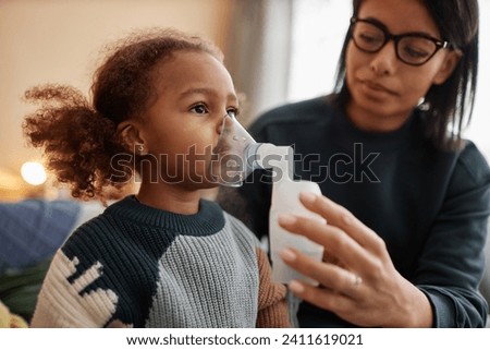 Medium close up shot of little African American girl inhaling medicine through nebulizer while defocused mom holding face mask on daughters face