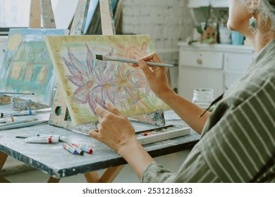 Medium close up of unrecognizable senior woman painting colorful big buds of flowers on white canvas during art therapy - Powered by Shutterstock