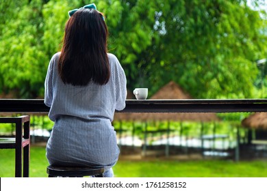 Medium Closes Up Back Of A Black Haired Young Woman In Pajamas Sitting Next To A Table Bar With A Cup Of Hot Coffee In Front Of Shelter Under The Bamboo Grove Blurred Background In The Morning.