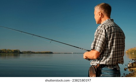 Medium close-up of an adult man in jeans and a shirt fishing on the banks of a picturesque river. A mature man with a short haircut and a little gray in his hair catches fish with a spinning rod - Powered by Shutterstock