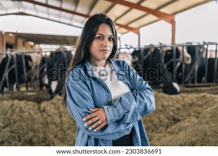 Medium close-up portrait of a young woman farmer looking at the camera with her arms crossed with her cows.