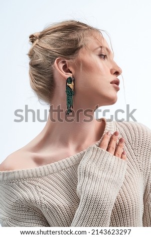 Medium close-up shot of a blond-haired lady in a white pullover with beaded earrings shaped as parrots. The lady is posing on the light background.