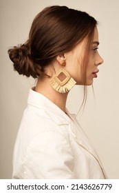 Medium close-up shot of a woman with boho rhombus earrings with a fringe. These earrings are decorated with raffia weaving. The woman in a white shirt with earrings is posing on a beige background.