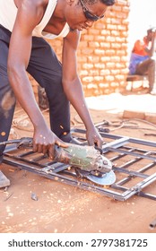 Medium close-up, side-angled shot of a Black man in his late twenties intensely grinding a steel frame with a handheld angle grinder, bright orange sparks illuminating dust in the air. He wears a white tank top, stained work trousers, and wraparound sunglasses, arms taut with focus. Background shows another man observing from a wooden bench against a sun-bleached brick wall, under bright daylight casting warm golden tones across the dry, reddish-brown earth scattered with metal shavings.