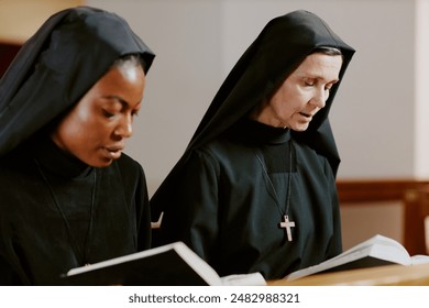 Medium closeup of two ethnically diverse young and mature Catholic nuns praying while sitting on pew