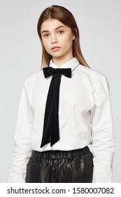 Medium Full Shot Of A Blonde Lady In A White Cotton Shirt With A Showy Black Velvet Necktie With Long Stripes. The Girl Is Wearing A Black Leather Bottom And Posing On The Light-gray Background.