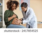 Medium full shot of senior Caucasian female gynecologist using hands for examining cheerful pregnant African American womans uncovered abdomen in clinic