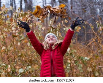 Medium Horizontal Photo Of Pretty Smiling Young Woman With Long Blond Hair Wearing Red Puffy Winter Coat Looking Up While Throwing Dry Leaves Over Her Head