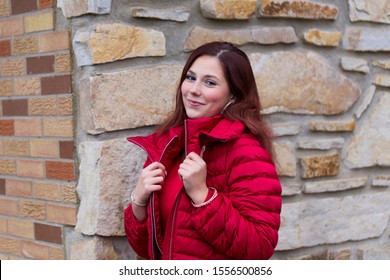 Medium Horizontal Photo Of Pretty Young Woman With Long Mahogany Hair Smiling Seductively And Wearing Red Dress And Puffy Winter Coat While Standing In Front Of Stone And Brick House