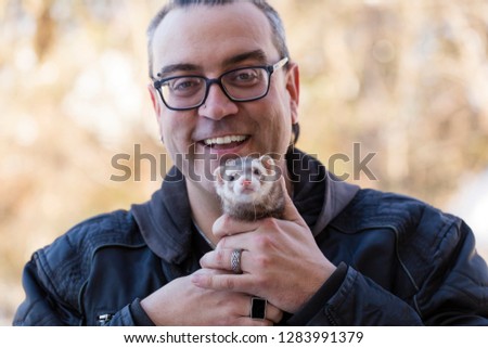 Medium horizontal portrait of ferret staring intently while held by smiling man with slicked back hair and soul patch wearing eyeglasses and a black coat against garden background