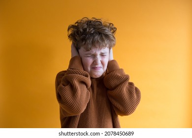 Medium horizontal shot of boy in rust sweater covering his ears with his hands to demonstrate loud noise and making a funny face  against orange background
